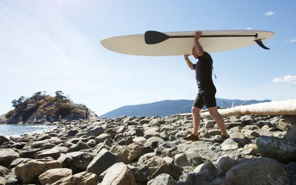 Mature adult carrying paddle board across the beach towards the sea.